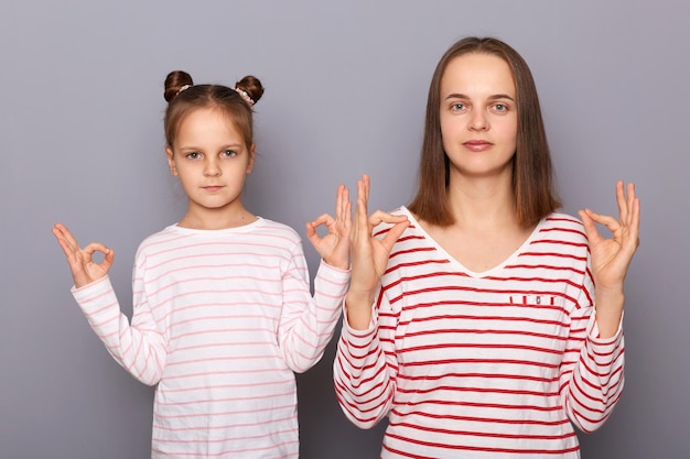 Portrait d'une mère calme et détendue sérieuse avec sa fille portant des vêtements décontractés isolés sur fond gris pratiquant le yoga ensemble se relaxant en regardant la caméra