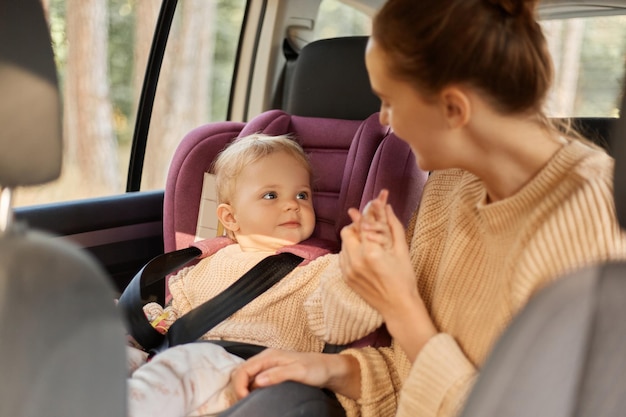 Portrait d'une mère caucasienne souriante en pull beige et de sa petite fille en siège bébé voyageant en voiture jouant ensemble pendant leur voyage exprimant le bonheur