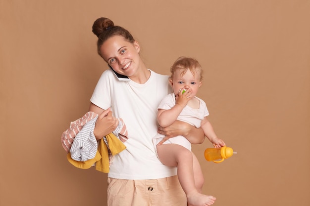 Portrait d'une mère occupée tenant une petite fille et des vêtements isolés sur fond marron femme portant un t-shirt blanc parlant sur un téléphone portable exprimant des émotions positives