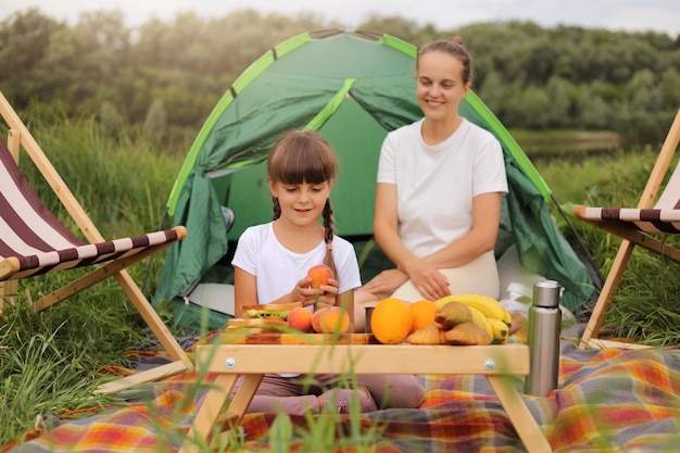 Portrait de mère et sa jolie fille avec des tresses en train de pique-niquer près de la rivière assis sur un plaid sur le sol des gens regardant la caméra en appréciant la nourriture savoureuse et l'air frais