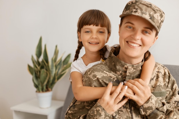 Portrait d'une mère souriante caucasienne et de son enfant femme portant un uniforme de camouflage et une casquette posant avec sa fille regardant la caméra avec un sourire heureux à pleines dents