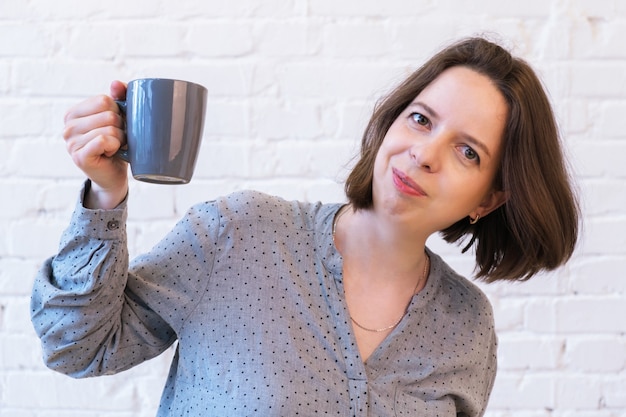 Portrait mi-long d'une jeune femme brune avec une tasse de café à la main sur fond de mur de briques blanches. La femme sourit et tient une tasse grise en céramique dans sa main.