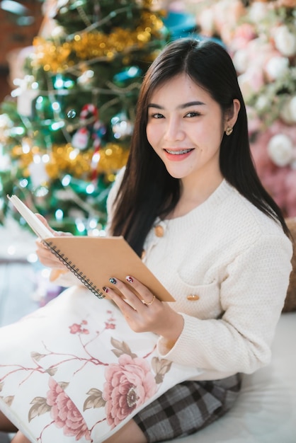 Portrait Mignon beau sourire positif jeune femme asiatique tenant de lire un livre à la maison dans le salon à l'intérieur Décoration Pendant les vacances de Noël et du Nouvel An.