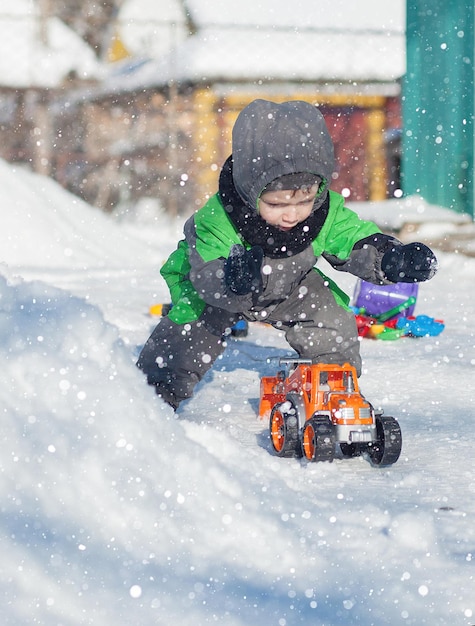 Portrait de mignon petit bambin assis sur la neige et jouant avec son jouet de tracteur jaune dans le parc Enfant jouant à l'extérieur Heureux garçon avec jouet de construction Concept de style de vie