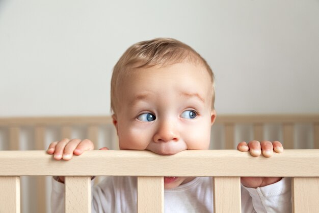 Portrait de mignon petit bébé aux yeux bleus en body blanc mordant la tête de lit en bois sur fond intérieur blanc, espace copie, horizontal