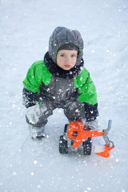 Portrait de mignon petit enfant assis sur la neige et jouant avec son jouet tracteur jaune dans le parc