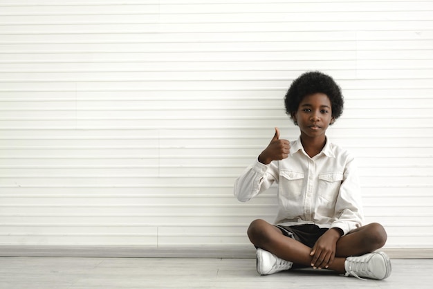 Portrait d'un mignon petit garçon afro-américain noir assis sur le sol avec un bon pouce vers le haut sur fond de mur blanc