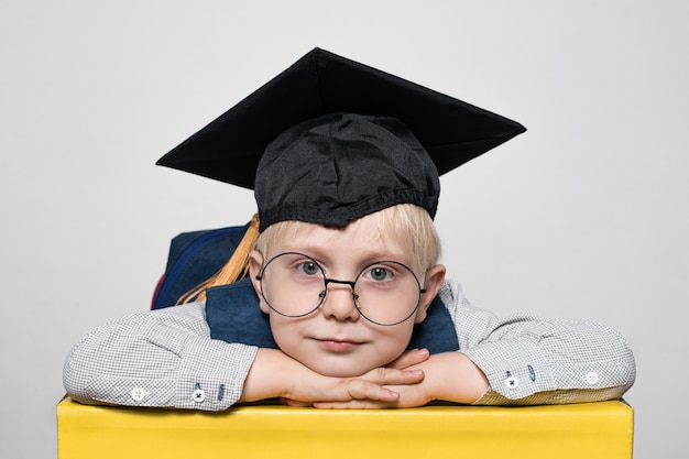 Portrait d'un mignon petit garçon blond dans de grandes lunettes, un chapeau académique et un sac à dos