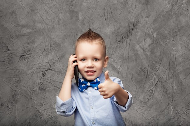 Portrait d'un mignon petit garçon en chemise bleue et noeud papillon avec téléphone mobile contre texture gris