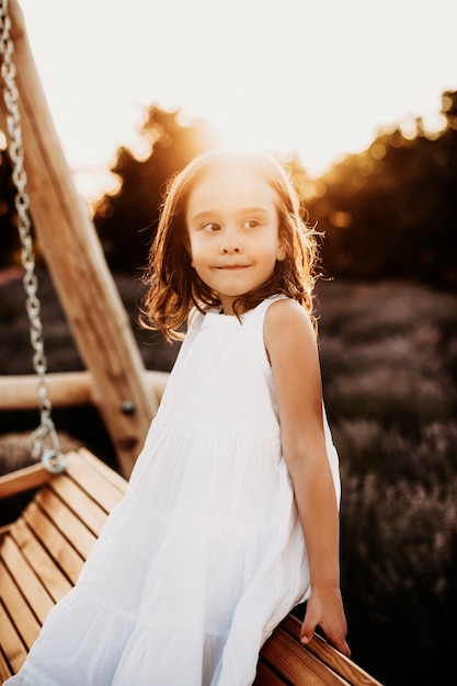 Portrait d'une mignonne petite fille assise sur une balançoire en bois à la recherche de suite souriant contre le coucher du soleil habillé en robe blanche.