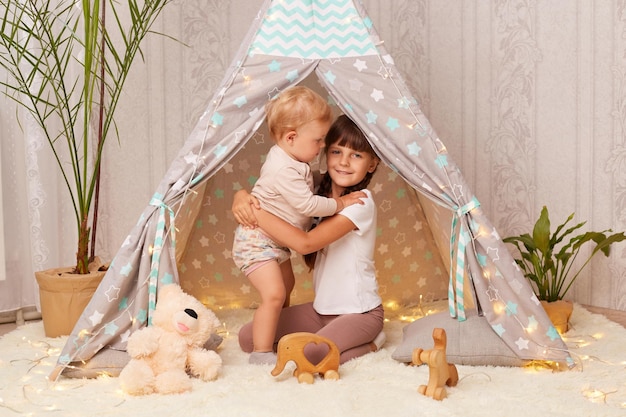 Portrait de mignonnes petites sœurs jouant dans un wigwam étreignant tout en jouant ensemble une fille aînée et un tout-petit bébé assis dans une tente peetee s'amusant avec intérêt