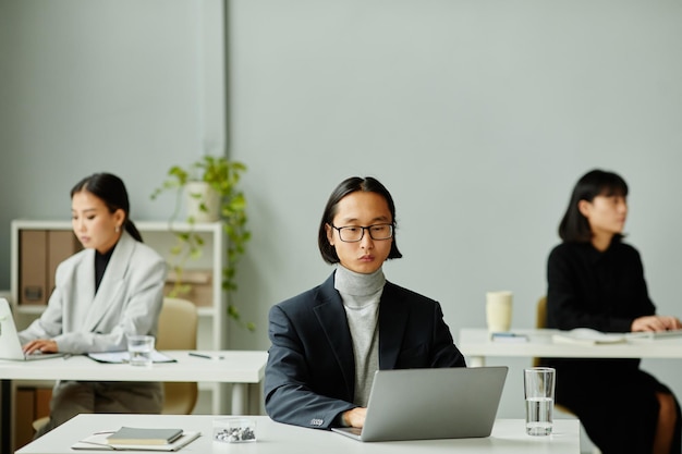 Portrait minimal d'un homme d'affaires asiatique utilisant un ordinateur portable avec des collègues féminines en arrière-plan, espace de copie