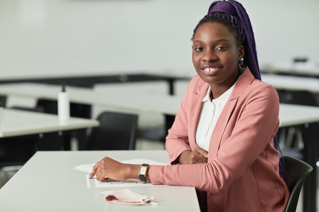 Portrait minimal d'une jeune femme afro-américaine souriante à la caméra à l'école alors qu'elle était assise au bureau dans la salle de classe, espace pour copie