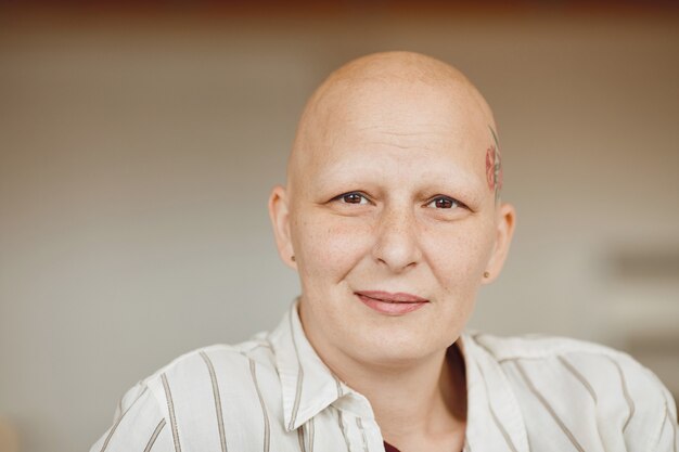 Portrait minimal de la tête et des épaules de femme adulte chauve regardant la caméra et souriant alors qu'il était assis sur un canapé dans un intérieur aux tons chauds, alopécie et sensibilisation au cancer, espace copie