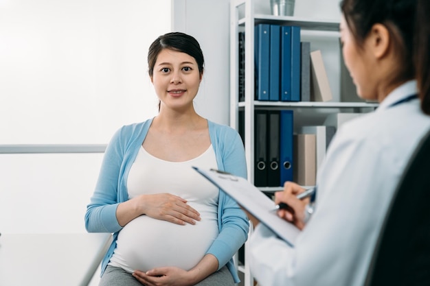 portrait de mise au point sélective d'une mère enceinte asiatique heureuse garde sa main sur le ventre et sourit à la caméra lors d'une visite régulière à son gynécologue à la clinique.