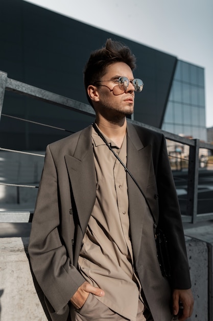 Portrait à la mode d'un beau jeune homme d'affaires professionnel avec des lunettes de soleil vintage en costume de mode gris sur fond urbain moderne au soleil