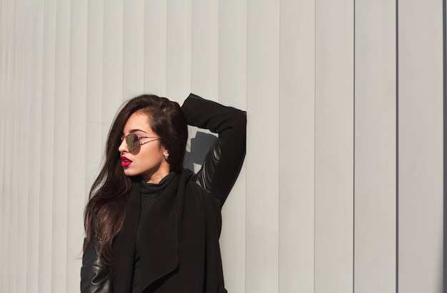 Portrait de mode d'un beau modèle aux cheveux longs en manteau tendance et lunettes élégantes posant dans le bâtiment