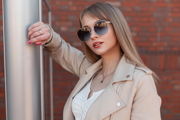 Portrait de mode féminine d'une belle jeune fille zoomer avec des lunettes de soleil rondes vintage et une veste en cuir blanche se tient près d'un stand de publicités métalliques dans la ville