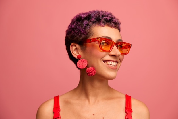 Portrait de mode d'une femme avec une coupe de cheveux courte dans des lunettes de soleil colorées avec des accessoires inhabituels avec des boucles d'oreilles souriantes sur un fond rose brillant Photo de haute qualité