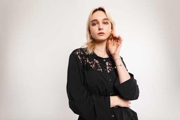 Portrait à la mode jeune femme blonde avec des boucles d'oreilles incroyables avec des lèvres sexy en robe noire à la mode avec de la dentelle près du mur vintage blanc