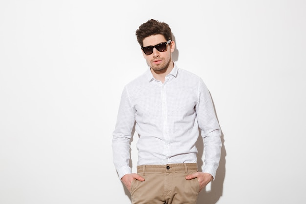 Portrait de mode jeune homme habillé en chemise et lunettes de soleil posant sur l'appareil photo avec les mains dans les poches, sur un espace blanc avec ombre