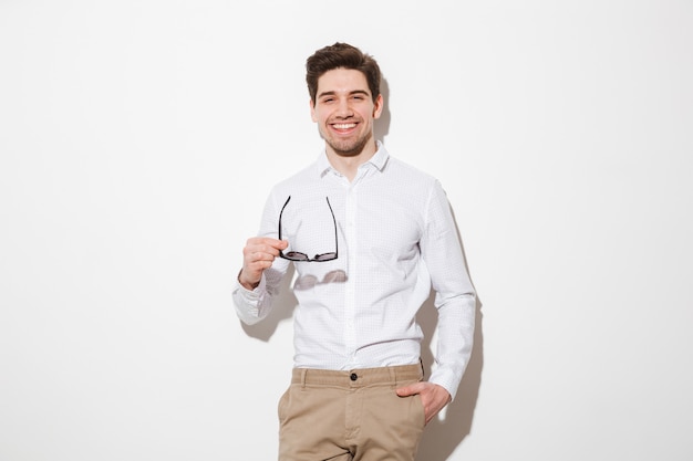 Portrait de mode jeune homme habillé en chemise souriant et posant sur l'appareil photo tenant des lunettes de soleil noires, sur un espace blanc avec ombre