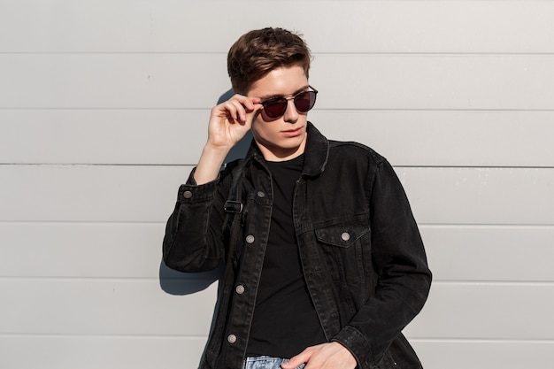 Portrait à la mode jeune homme à la mode dans une veste noire en denim élégante dans des lunettes de soleil vintage