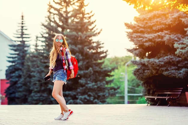 Portrait de mode d'une jeune jolie femme hipster en plein air avec de longs cheveux et un sac à dos rouge sous le soleil