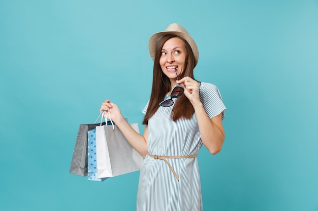 Portrait à la mode jolie femme heureuse en robe d'été, chapeau de paille, lunettes de soleil tenant des sacs de paquets avec des achats après le shopping isolé sur fond bleu pastel. Copiez l'espace pour la publicité.