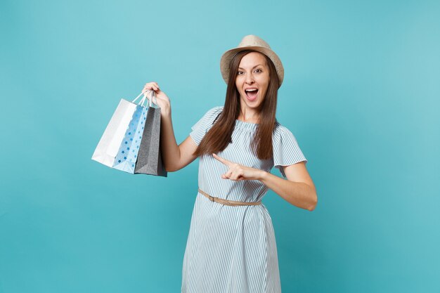 Portrait à la mode souriante belle femme caucasienne en robe d'été, chapeau de paille tenant des sacs de paquets avec des achats après le shopping isolé sur fond bleu pastel. Copiez l'espace pour la publicité.