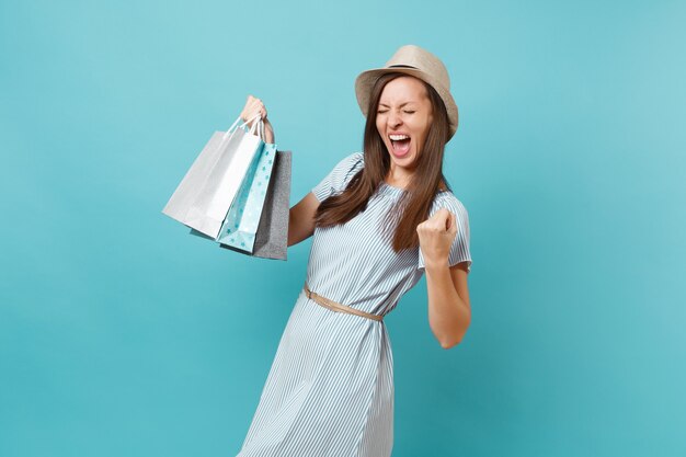 Portrait à la mode souriante belle femme caucasienne en robe d'été, chapeau de paille tenant des sacs de paquets avec des achats après le shopping isolé sur fond bleu pastel. Copiez l'espace pour la publicité.