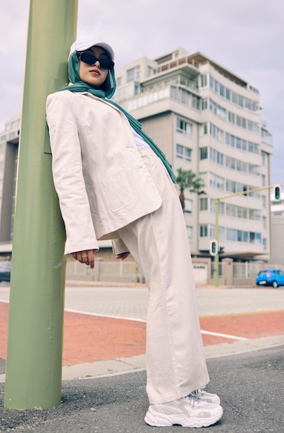 Portrait mode urbaine ou religion avec une femme musulmane appuyée contre un poteau dans une casquette et une écharpe pour la foi islamique de style contemporain ou le hijab avec un jeune arabe branché à l'extérieur dans une ville urbaine