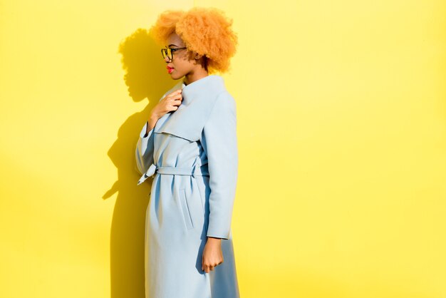 Photo portrait de mode de vie d'une belle femme africaine en manteau bleu debout sur le fond jaune. notion d'automne