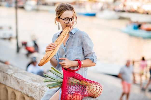 Portrait de mode de vie d'une jeune femme avec un sac en filet rempli de produits frais mangeant de la baguette en plein air dans la vieille ville