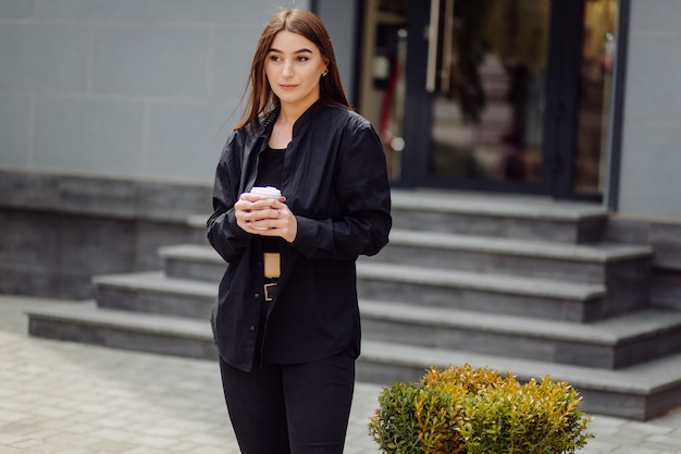 Portrait de mode de vie en plein air d'une superbe fille brune. Boire du café et marcher dans la rue de la ville.