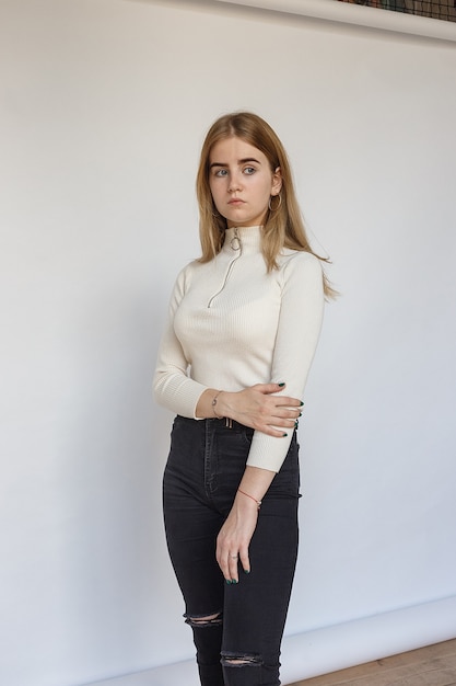 Portrait d'un modèle adolescent réfléchi portant un pull blanc et un jean noir. Caucasien maigre jeune femme aux cheveux blonds se tient devant le mur de papier blanc. jolie dame naturelle debout
