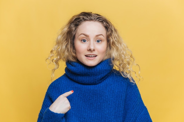 Portrait d'un modèle féminin attrayant surpris avec des cheveux bouclés indique à l'espace de copie de son nouveau pull d'hiver chaud montre de l'espace pour votre publicité ou votre conception isolée sur fond jaune