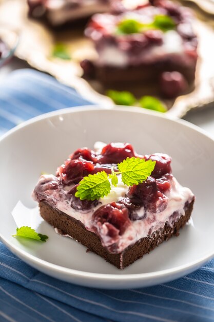 Portrait d'un morceau de brownie avec une crème, des cerises et des feuilles de menthe fraîche sur une assiette.