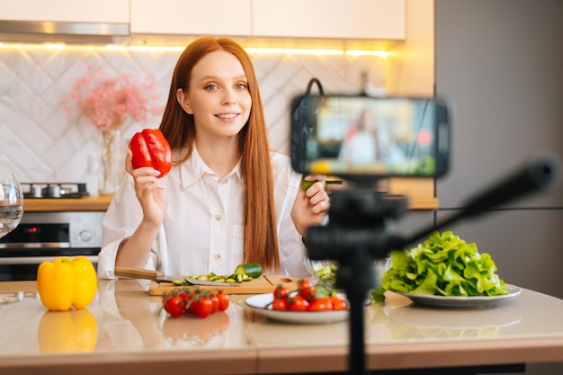 Portrait moyen d'une jeune blogueuse rousse joyeuse tenant à la main des poivrons rouges enregistrant une vidéo de tutoriel en direct sur une alimentation végétarienne saine sur la caméra d'un téléphone portable dans la cuisine
