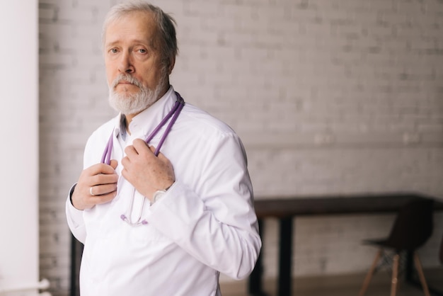 Portrait moyen d'un médecin de sexe masculin mûr et confiant en blouse blanche et stéthoscope debout dans une salle de bureau d'hôpital lumineuse. Médecin adulte professionnel avec barbe blanche et cheveux gris sur le lieu de travail.