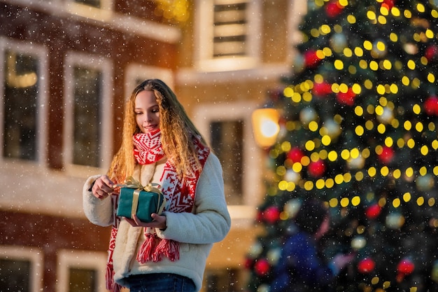 Portrait de Noël d'une belle fille vêtue de vêtements chauds et d'une écharpe debout dans la rue décorée pour le nouvel an sur fond d'arbre de Noël avec un cadeau dans les mains.