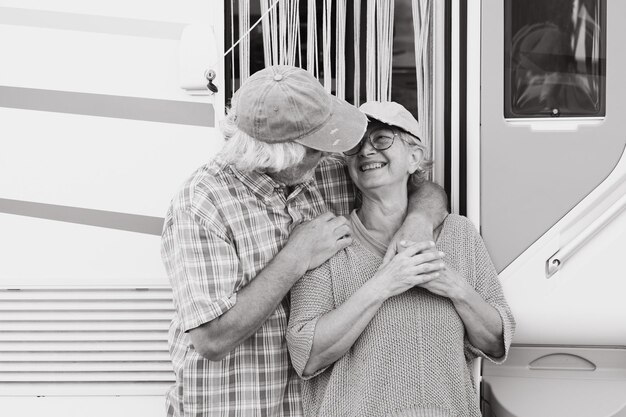 Portrait noir et blanc d'un couple de personnes âgées joyeux en voyage d'agrément debout à l'extérieur d'un camping-car se serrant dans ses bras et se regardant