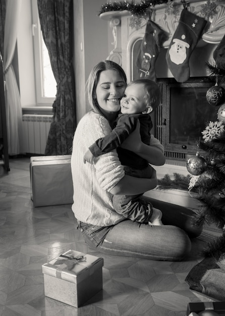 Portrait noir et blanc d'une jeune mère heureuse serrant son petit garçon de 1 an à l'arbre de Noël