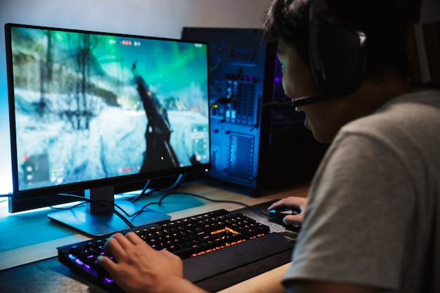 Portrait of Asian happy gamer boy jouer à des jeux vidéo en ligne sur ordinateur dans une pièce sombre, portant des écouteurs avec microphone et à l'aide d'un clavier coloré rétroéclairé