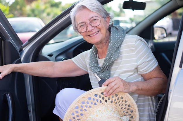 Portrait of attractive smiling senior woman casual habillé entrant dans sa voiture tenant les clés et son chapeau looking at camera