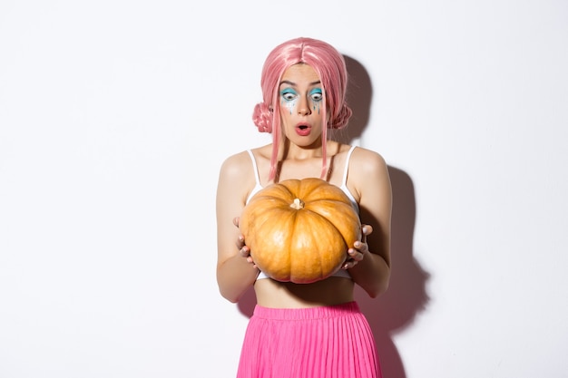 Portrait of attractive young party girl in pink wig regardant étonné à la citrouille, célébrant l'halloween, debout sur fond blanc