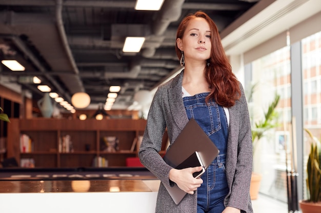 Portrait of Casually Dressed Young Businesswoman Standing In Modern Open Plan Workplace