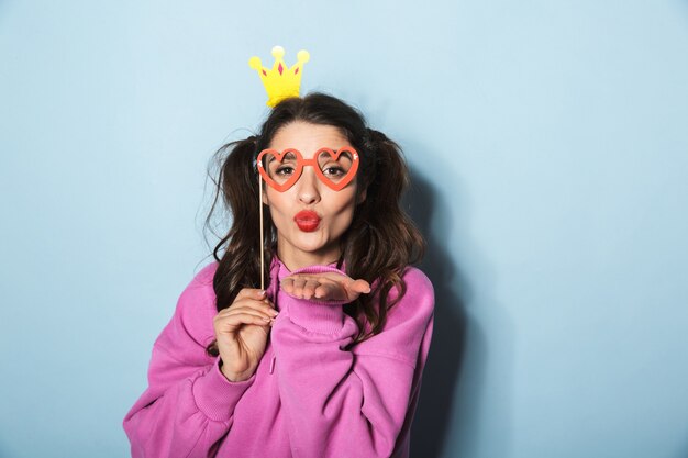 Portrait of cheerful princess girl wearing paper crown rire et tenant des lunettes de soleil drôles sur bâton sur bleu en studio