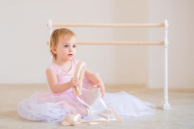 Portrait of cute blueeyed baby ballerine en rose assis près de la barre de ballet mettant des chaussures de pointe