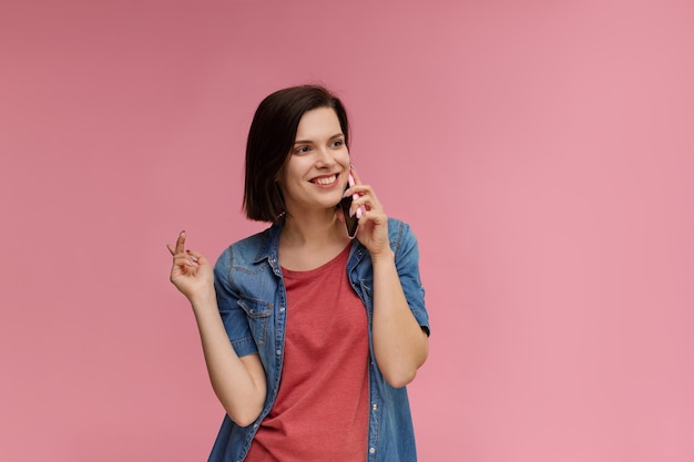 Portrait of cute happy brunette woman wearing t-shirt et jeans shirt parler sur téléphone mobile et souriant isolé sur fond rose